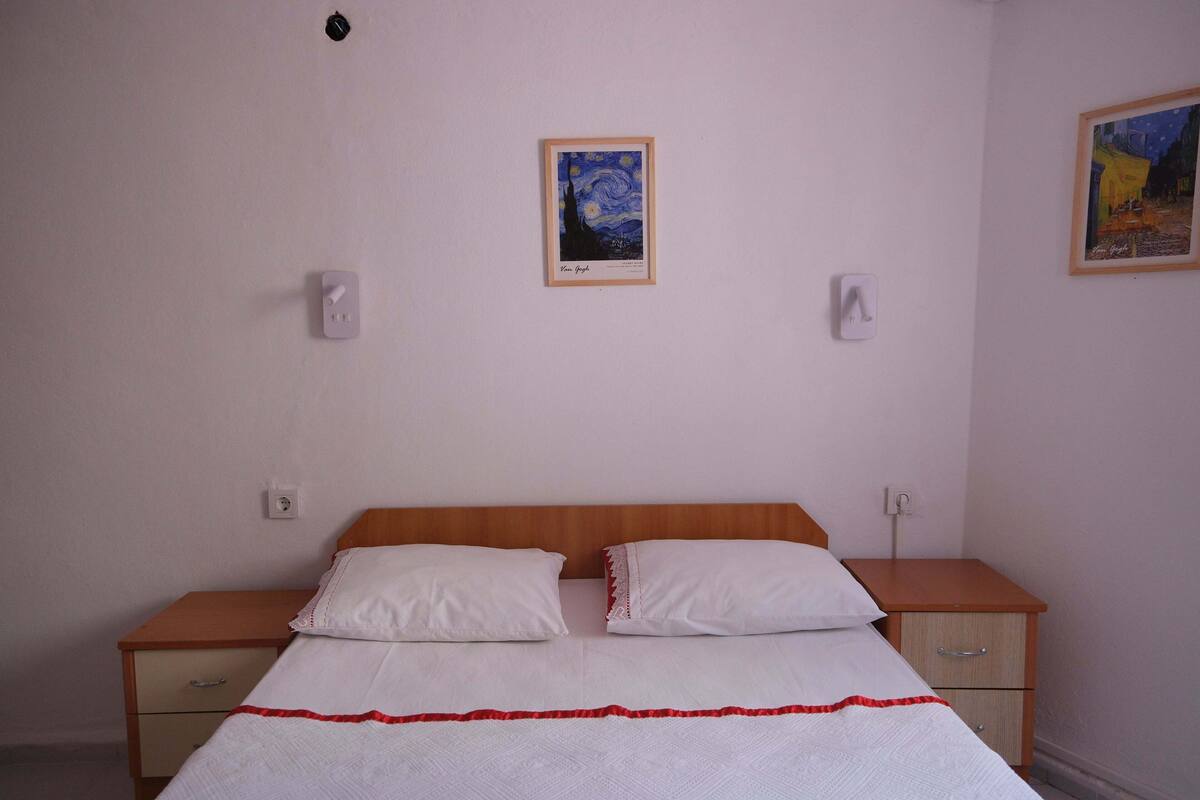 Un dormitorio con cabecera de madera, dos almohadas blancas, una colcha blanca con detalles rojos, una mesita de noche de madera con cajones y dos interruptores de luz en la pared.