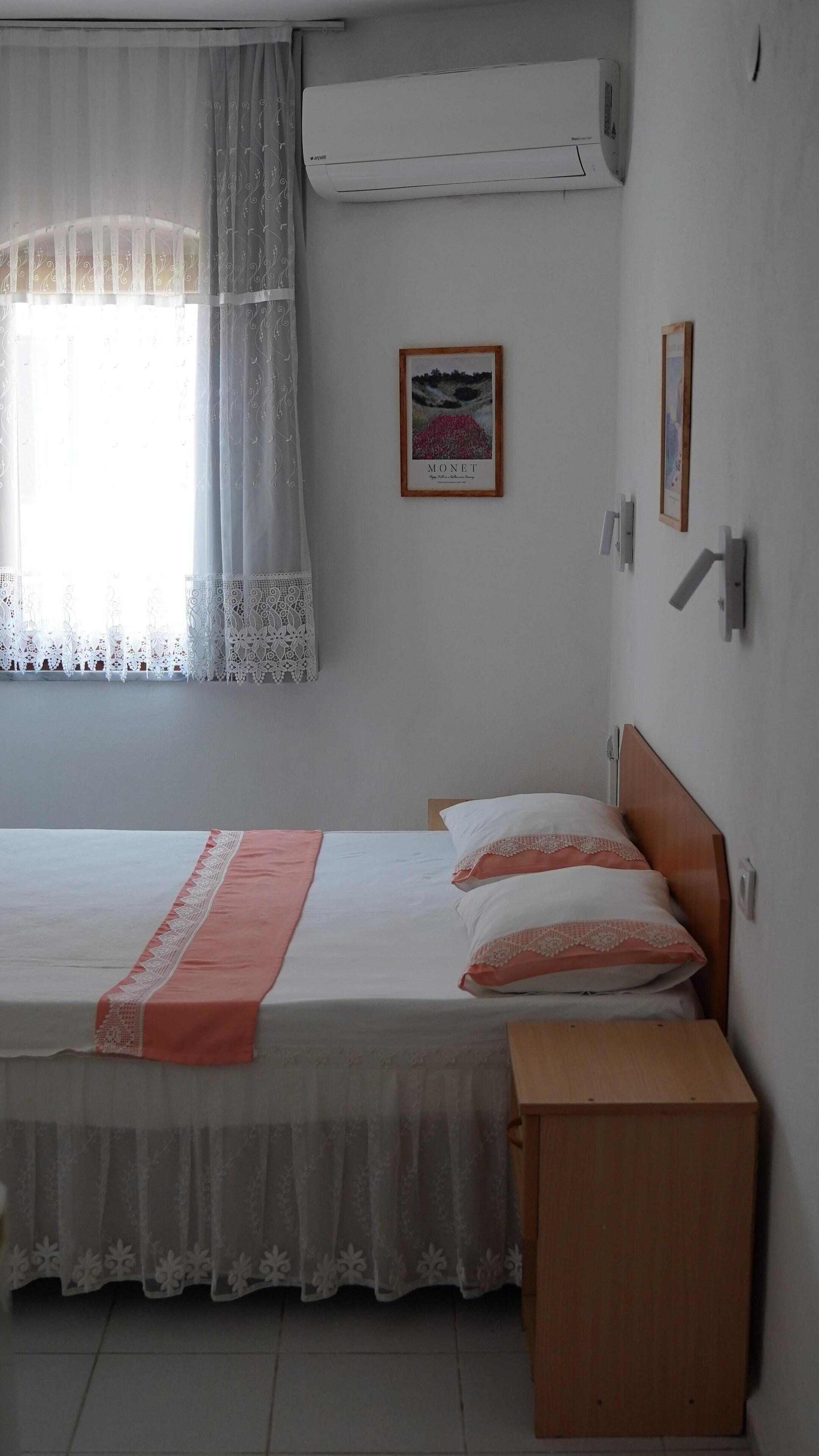 Un dormitorio con cama, mesita de noche, aire acondicionado y ventana con cortinas.