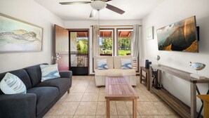 A living room with a sofa, a coffee table, a TV, and a ceiling fan. - NEW! Village Manor D21, Distant Ocean Views, Walk to Kapaa Town and Beach (Kapaa)