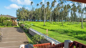 A view from a covered area overlooking a parking lot and a green field with palm trees. - NEW! Village Manor D21, Distant Ocean Views, Walk to Kapaa Town and Beach (Kapaa)