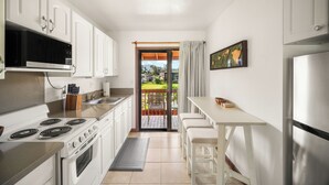 A modern kitchen with white cabinets, a stove, microwave, and a refrigerator. There is a dining area with a table and chairs, and a view of a garden through a glass door. - NEW! Village Manor D21, Distant Ocean Views, Walk to Kapaa Town and Beach (Kapaa)