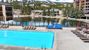 A swimming pool with lounge chairs and umbrellas, a view of a lake, and buildings in the background with snow-capped mountains. - On Main St. Hot Tubs, Easy Access to Peak 9. (Breckenridge)