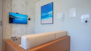 A modern hotel room with a large flat-screen TV mounted on the wall, a comfortable sofa, a marble accent wall, and a framed picture of a beach scene. - Elegance Residence (Bombinhas)