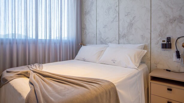 A neatly made bed with a beige blanket, a white pillow with a logo, and a nightstand with a phone and charger. - Elegance Residence (Bombinhas)