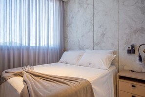 A neatly made bed with a beige blanket, a white pillow with a logo, and a nightstand with a phone and charger. - Elegance Residence (Bombinhas)