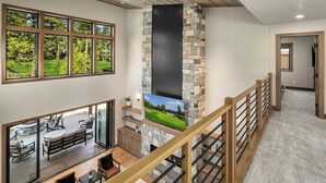 Un amplio salón con chimenea de piedra, grandes ventanales que ofrecen vistas a un paisaje verde y una escalera de madera con barandilla.