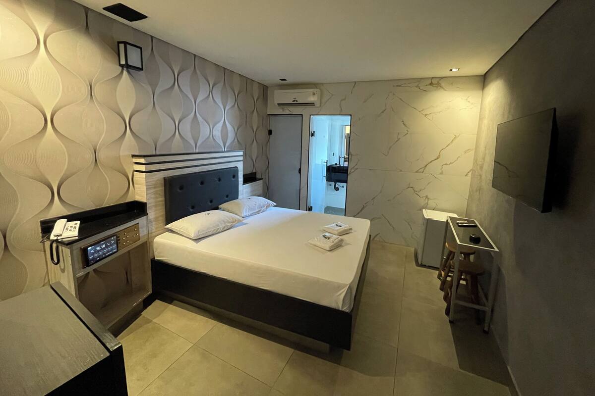 A modern hotel room with a large bed, a bedside table, a TV, and a bathroom visible through an open door.