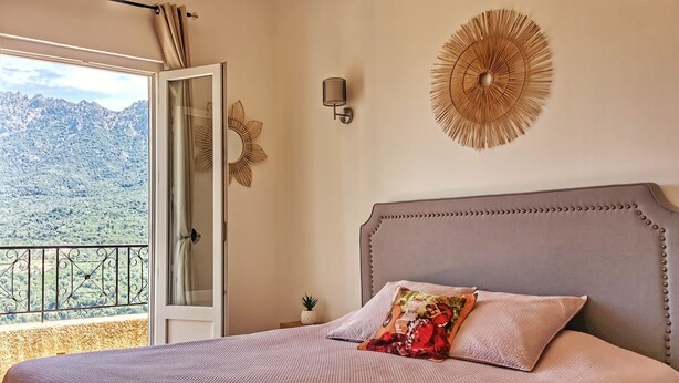 A bedroom with a bed, a view of mountains through an open door, a decorative wall piece, and a small plant.