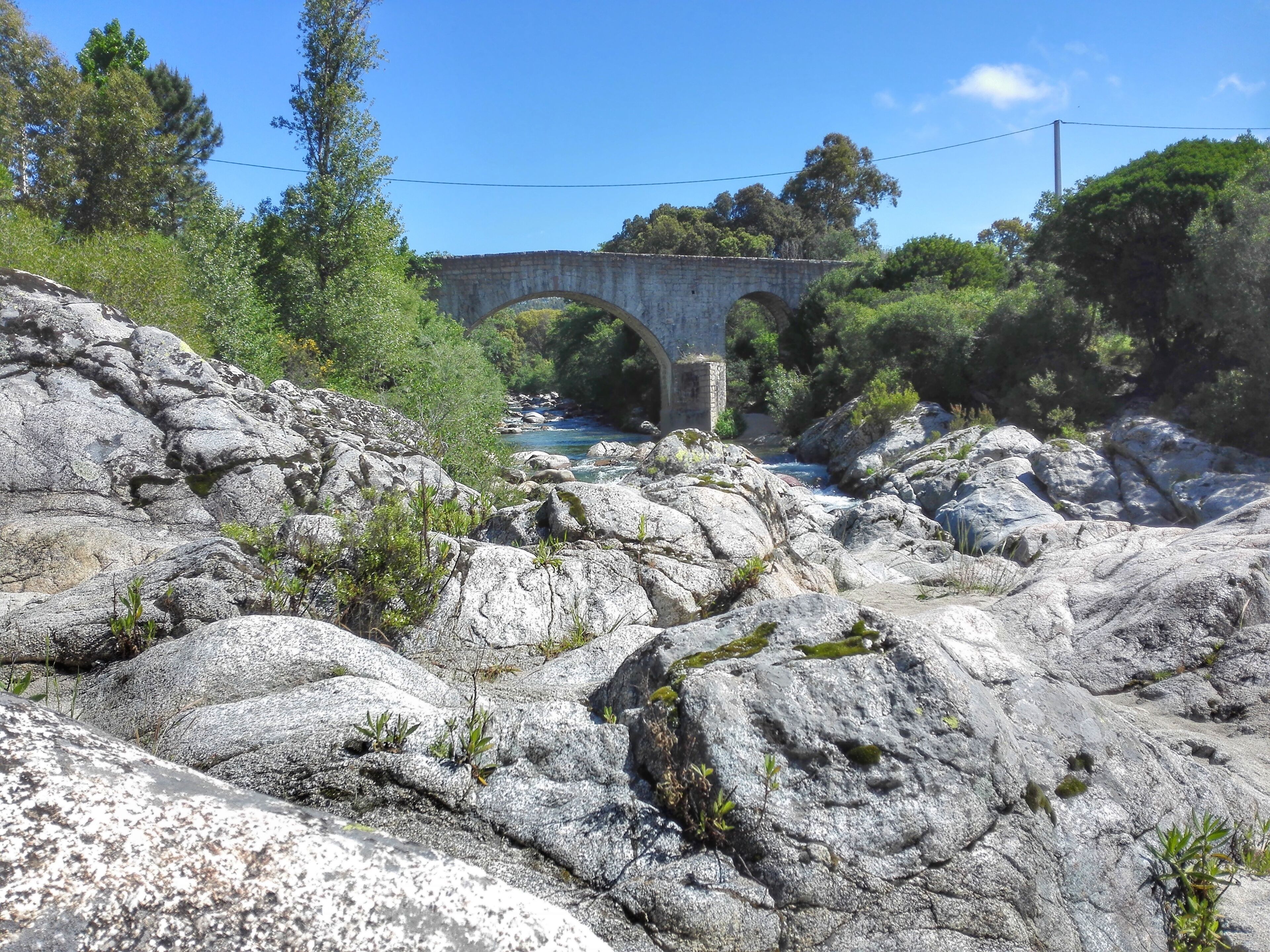 Un puente de piedra sobre un río, con terreno rocoso y vegetación circundante.