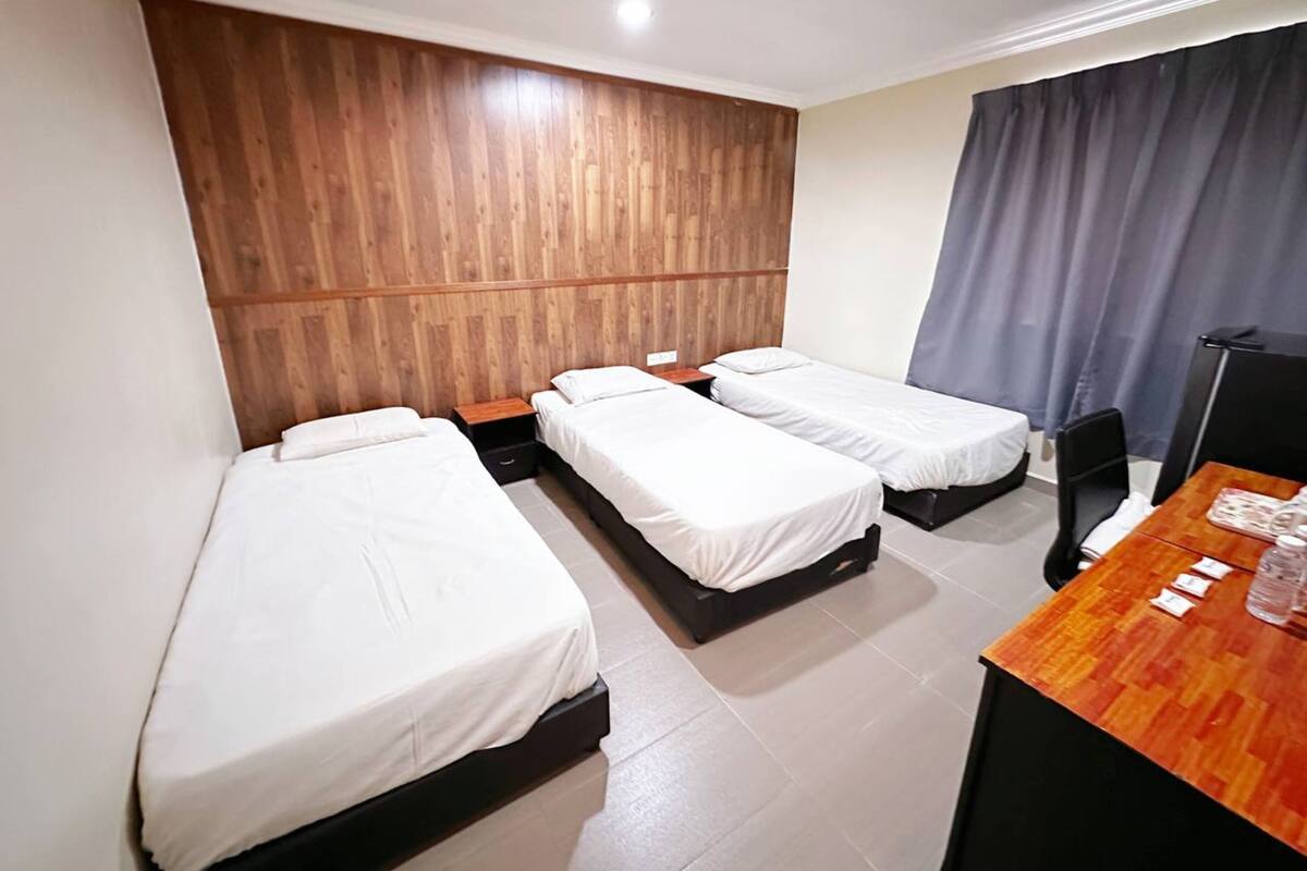 Ein Hotelzimmer mit drei Einzelbetten, einem hölzernen Kopfteil, einem Schreibtisch mit Stuhl und einem Vorhang.