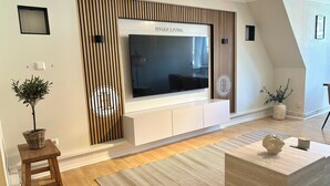 A modern living room with a large flat-screen TV mounted on a wall with a wooden panel design. There is a small wooden stool with a plant, a woven basket, and a textured rug on the floor. - HyggeLiving | 94m² | view of Marktkirche | OSCAR (Goslar)