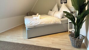 A neatly made bed with white linens and pillows, a grey sofa, a large green plant in a wicker basket, and a framed picture on the wall. - HyggeLiving | 94m² | view of Marktkirche | OSCAR (Goslar)