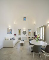 Una sala de estar moderna con un sofá blanco, una mesa de centro redonda y una mesa de comedor con sillas. Se observan obras de arte enmarcadas en las paredes, un radiador y una ventana con vistas.