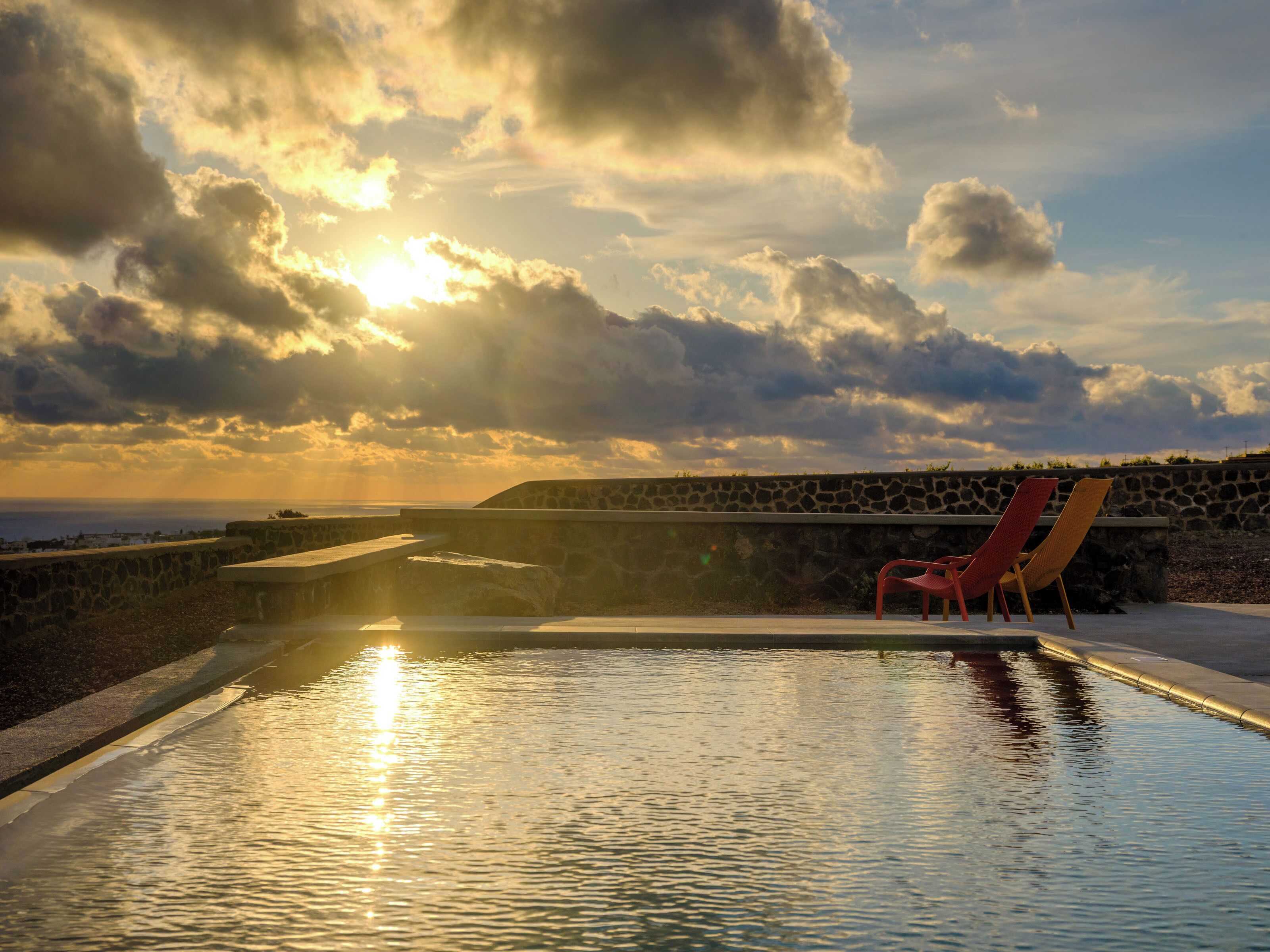 A pool with a stone wall and a red chair, overlooking a body of water and a distant view.