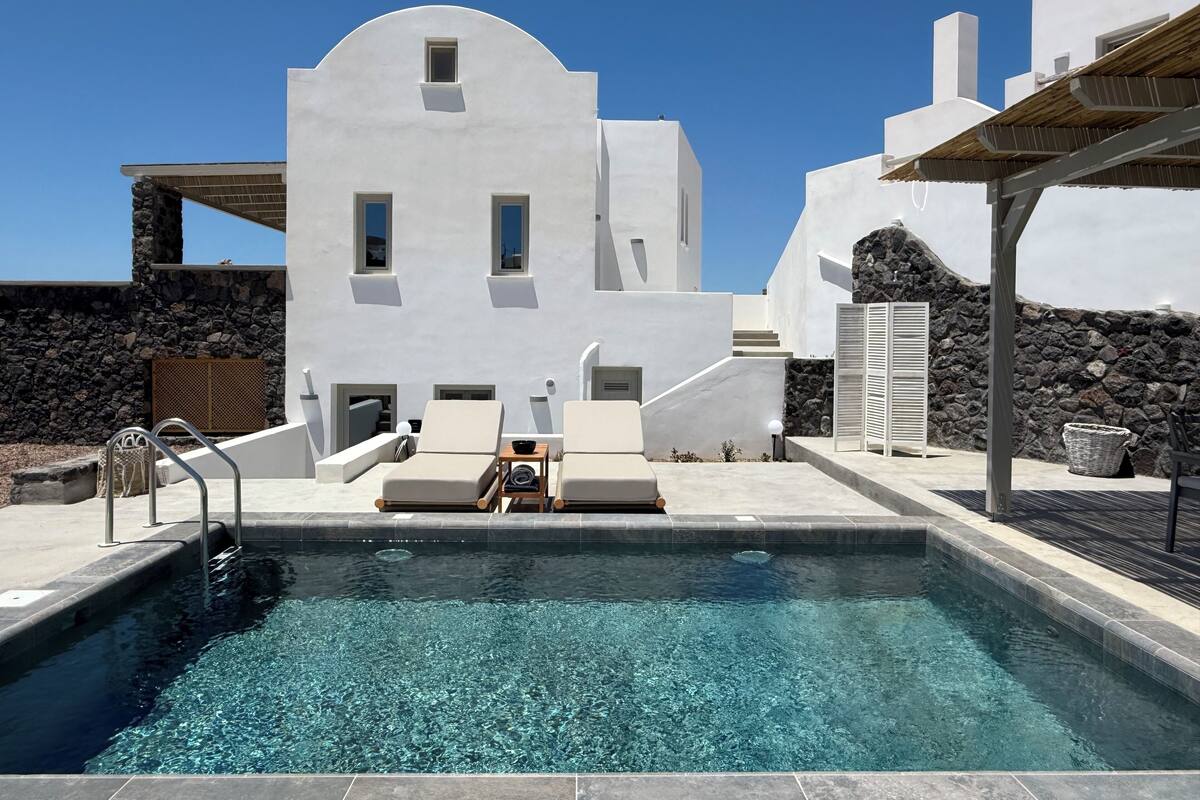 Un espace piscine moderne avec une eau d’un bleu limpide, des bâtiments blancs et des murs en pierre.