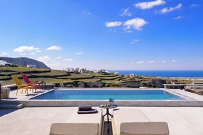 A poolside area with lounge chairs, a table with a glass, and a view of the sea and buildings. - Avan Luxury Villas (Santorini)