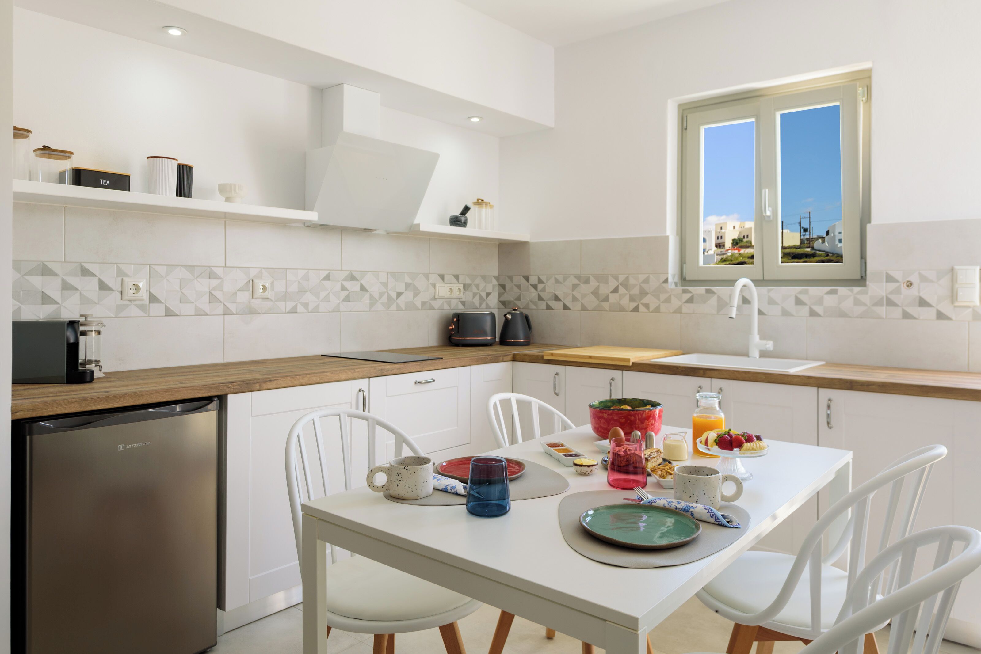 A modern kitchen with white cabinetry, a dishwasher, a window with a view, and a breakfast table set with plates, cups, and a fruit bowl.