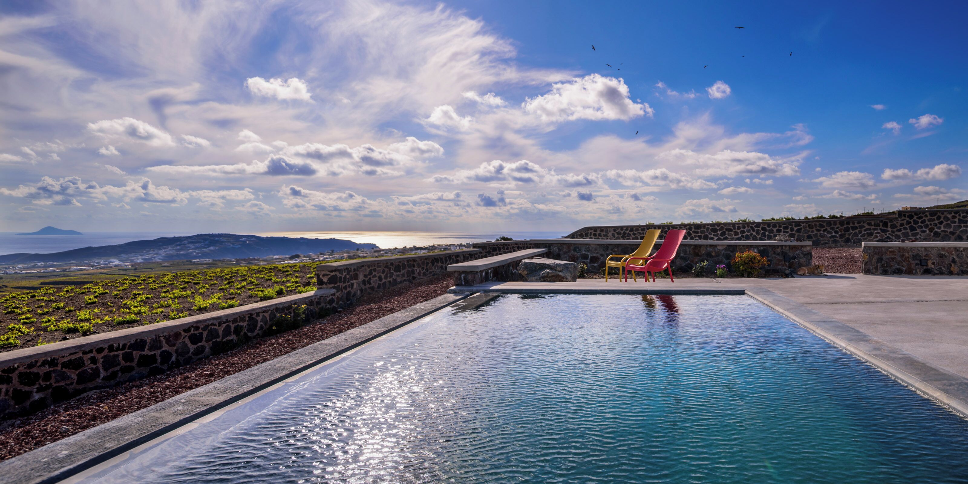 A swimming pool with a stone wall and colorful chairs, overlooking a landscape with mountains and a body of water.