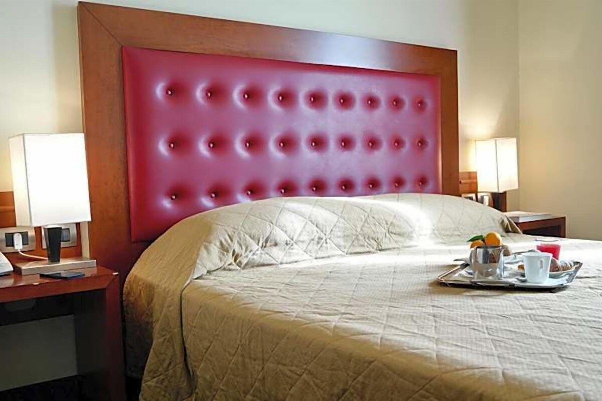 Une chambre d’hôtel avec un grand lit, une tête de lit rouge, des lampes de chevet, un téléphone et un plateau avec des articles pour le petit-déjeuner.