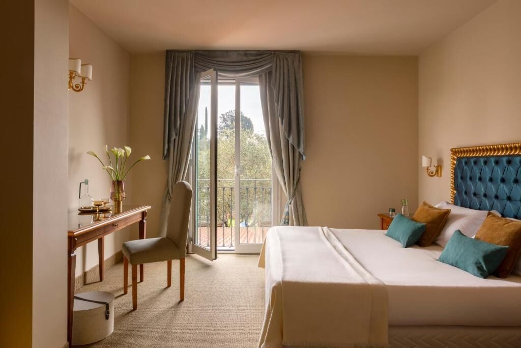 A hotel room with a bed, a desk, and a chair. There is a window with curtains, a view of the outdoors, and a wall-mounted lamp.
