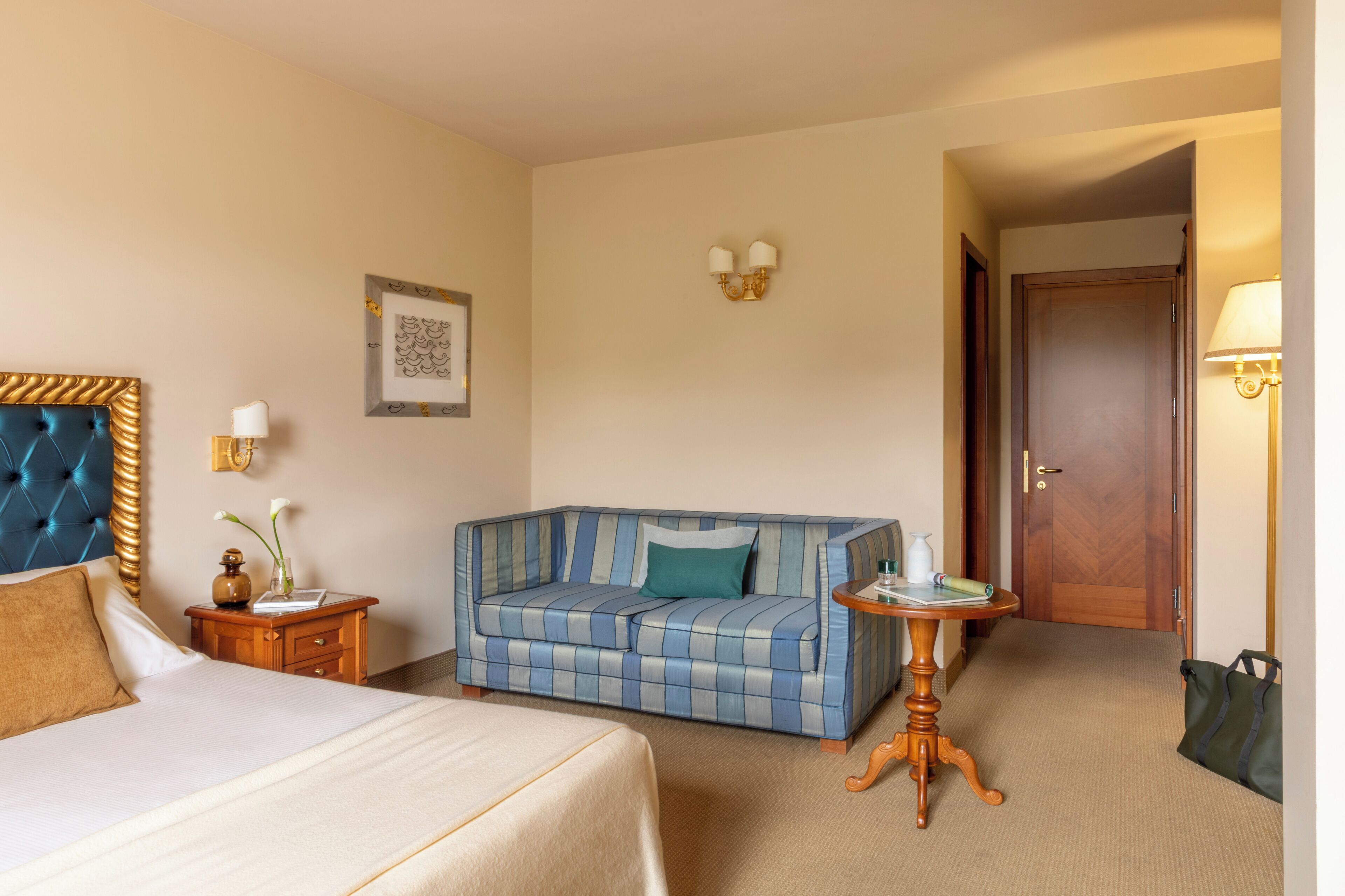 Una camera d'albergo con un letto, un divano blu a righe, un piccolo tavolino, un lampadario a parete e una porta.
