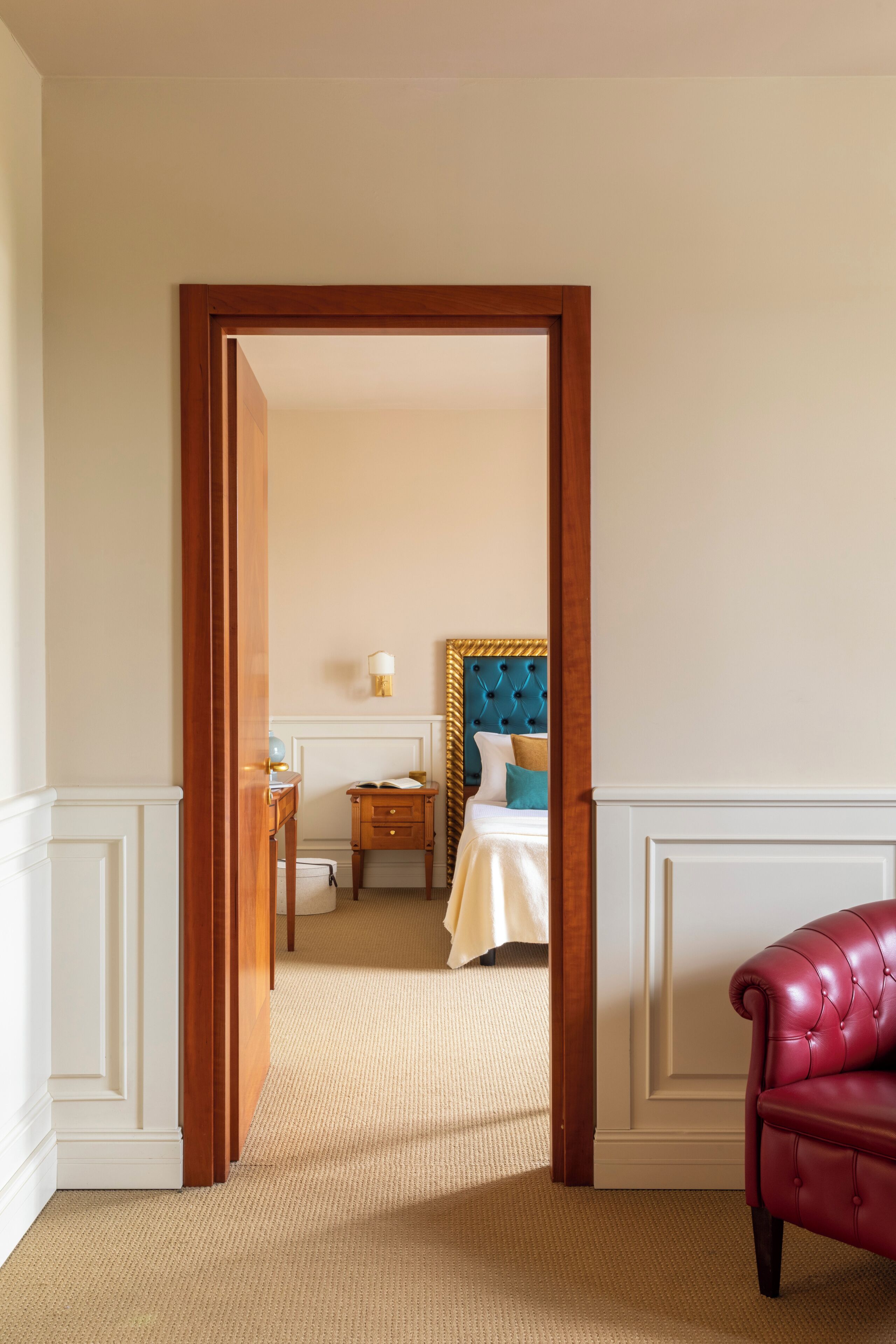 Une chambre d’hôtel avec une porte en bois, un lit doté d’une tête de lit capitonnée, une table de chevet et un fauteuil rouge.