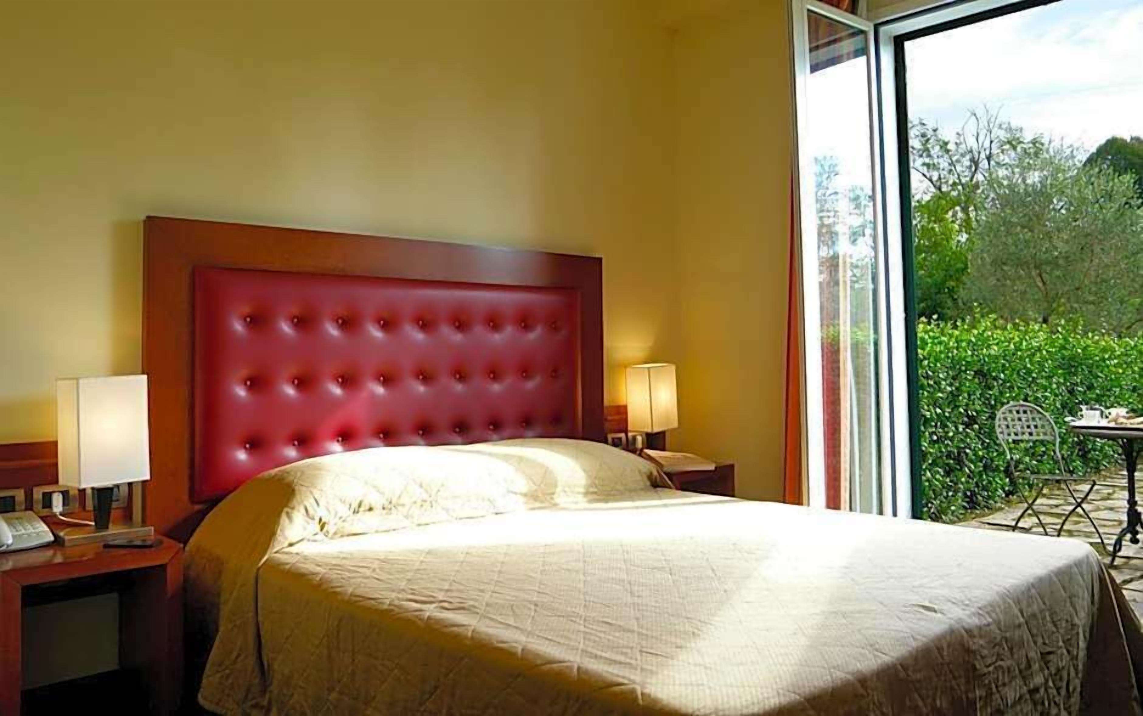 A hotel room with a large bed, two bedside tables, a window with a view of greenery, and a small table with a chair outside.