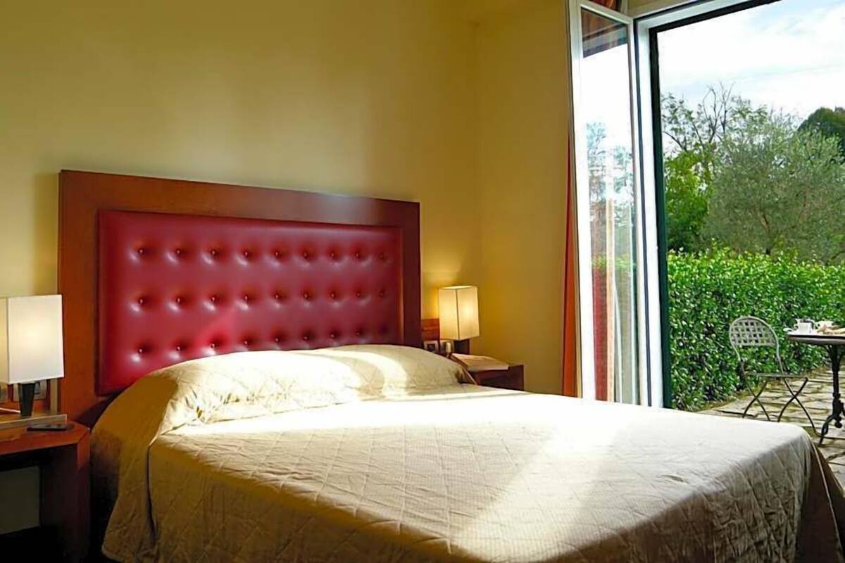 A hotel room with a large bed, two bedside tables, a window with a view of greenery, and a small table with a chair outside.