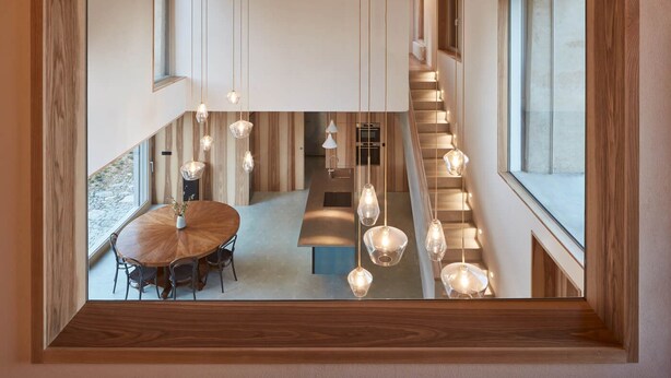 A modern interior with a wooden ceiling, a dining area with a round table and chairs, and a staircase with hanging pendant lights.