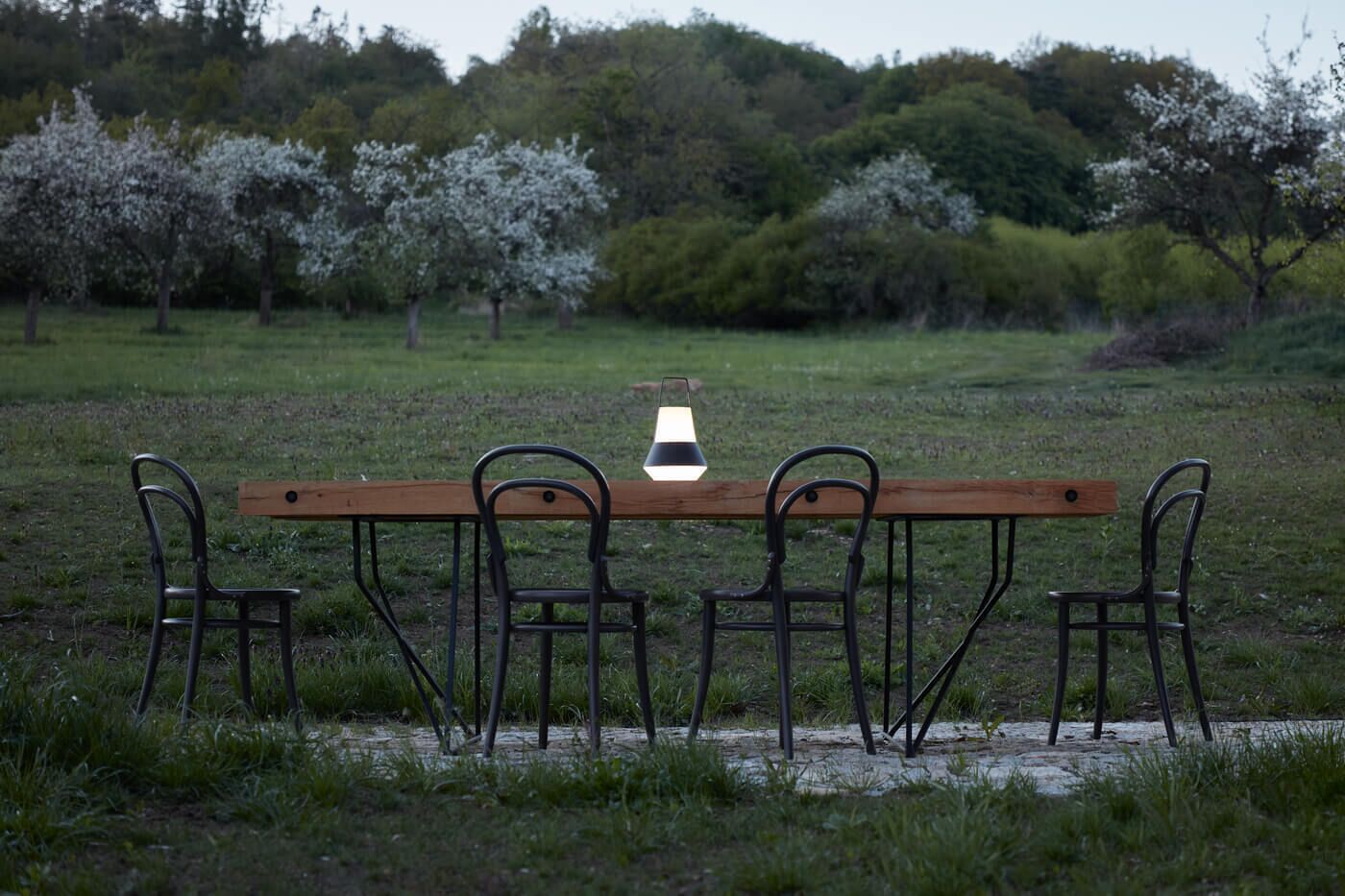 Un banco de madera con sillas de metal en un campo cubierto de hierba, con árboles al fondo.