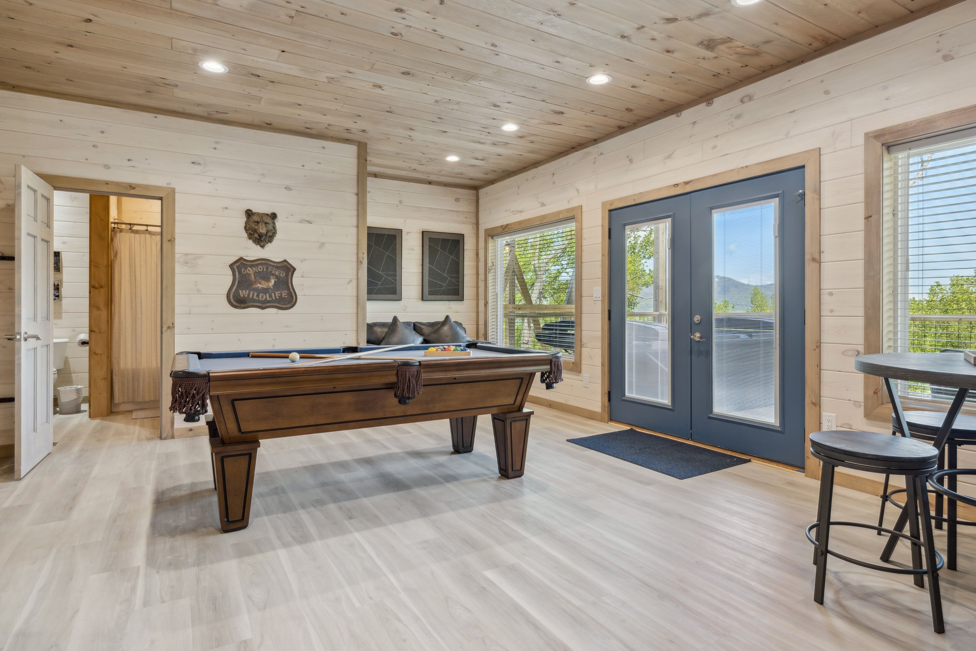 A pool table in a room with wooden walls and ceiling, a door, and a window with blinds.