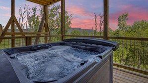 Uma banheira de hidromassagem com deck de madeira e vista panorâmica para árvores e montanhas ao pôr do sol.