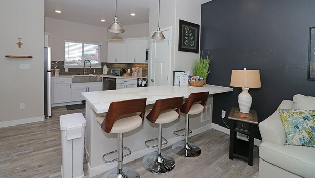 A modern kitchen with white cabinets, a central island with bar stools, and a dining area with a lamp and a couch. - Modern Ascend 2 Bedroom Townhouse (Grand Junction)