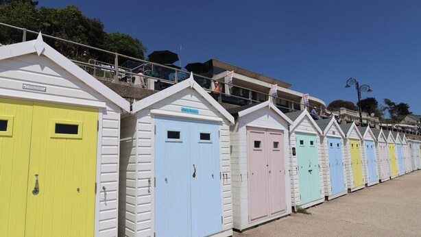 A row of colorful beach huts with white exteriors and pastel-colored doors, each with a small window and a door handle. - Bentley Rise (Lyme Regis)