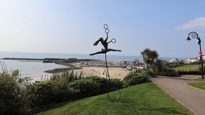 A coastal view with a sculpture of a figure balancing rings, a beach, and a pathway leading to a town. - Bentley Rise (Lyme Regis)