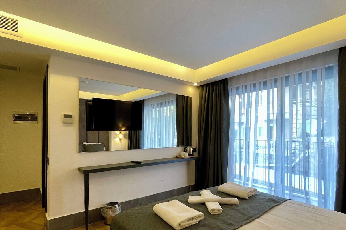 A hotel room with a bed, a bedside table, a mirror, a window with curtains, and a view of the outside.