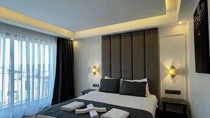 A modern hotel room with a large bed, two bedside tables, a wall-mounted light, and a view through sheer curtains. - Brow City Kaş (Kas)