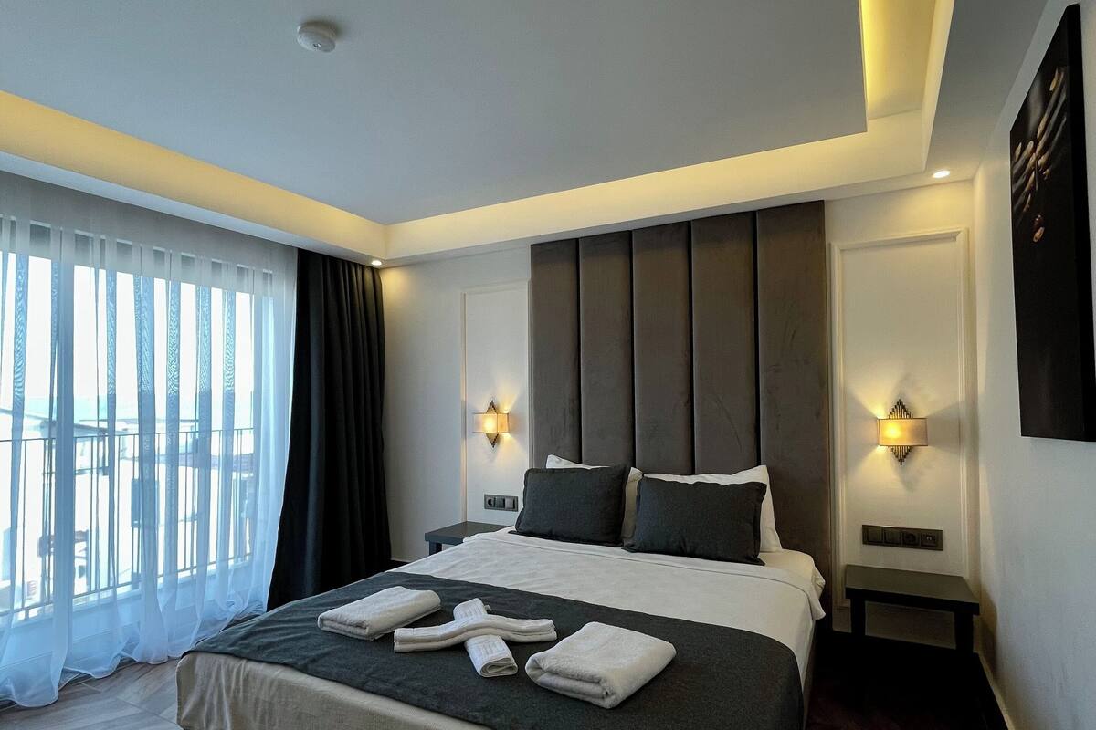 A modern hotel room with a large bed, two bedside tables, a wall-mounted light, and a view through sheer curtains.