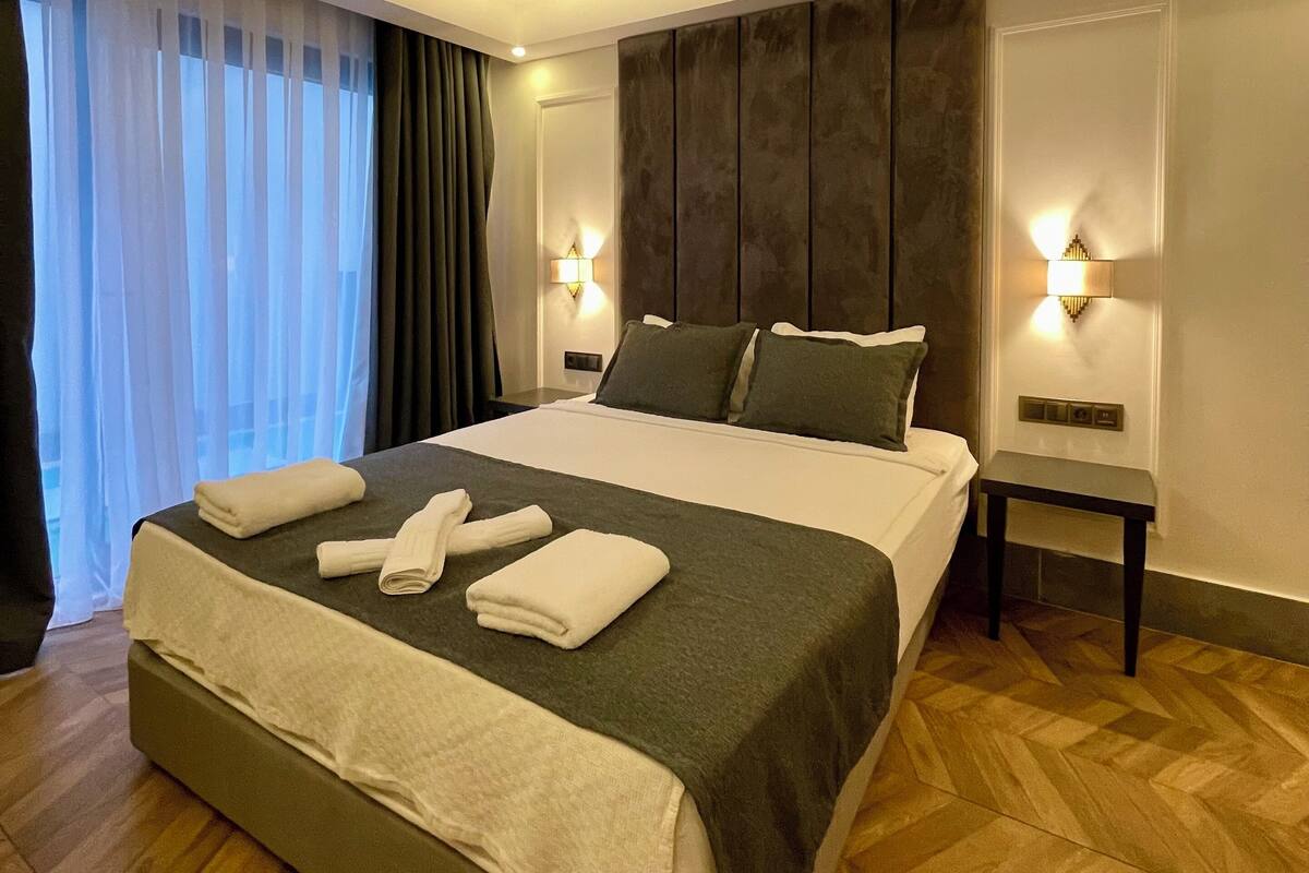 A modern hotel room with a large bed, two bedside tables with lamps, a wooden floor, and a view of the city through sheer curtains.
