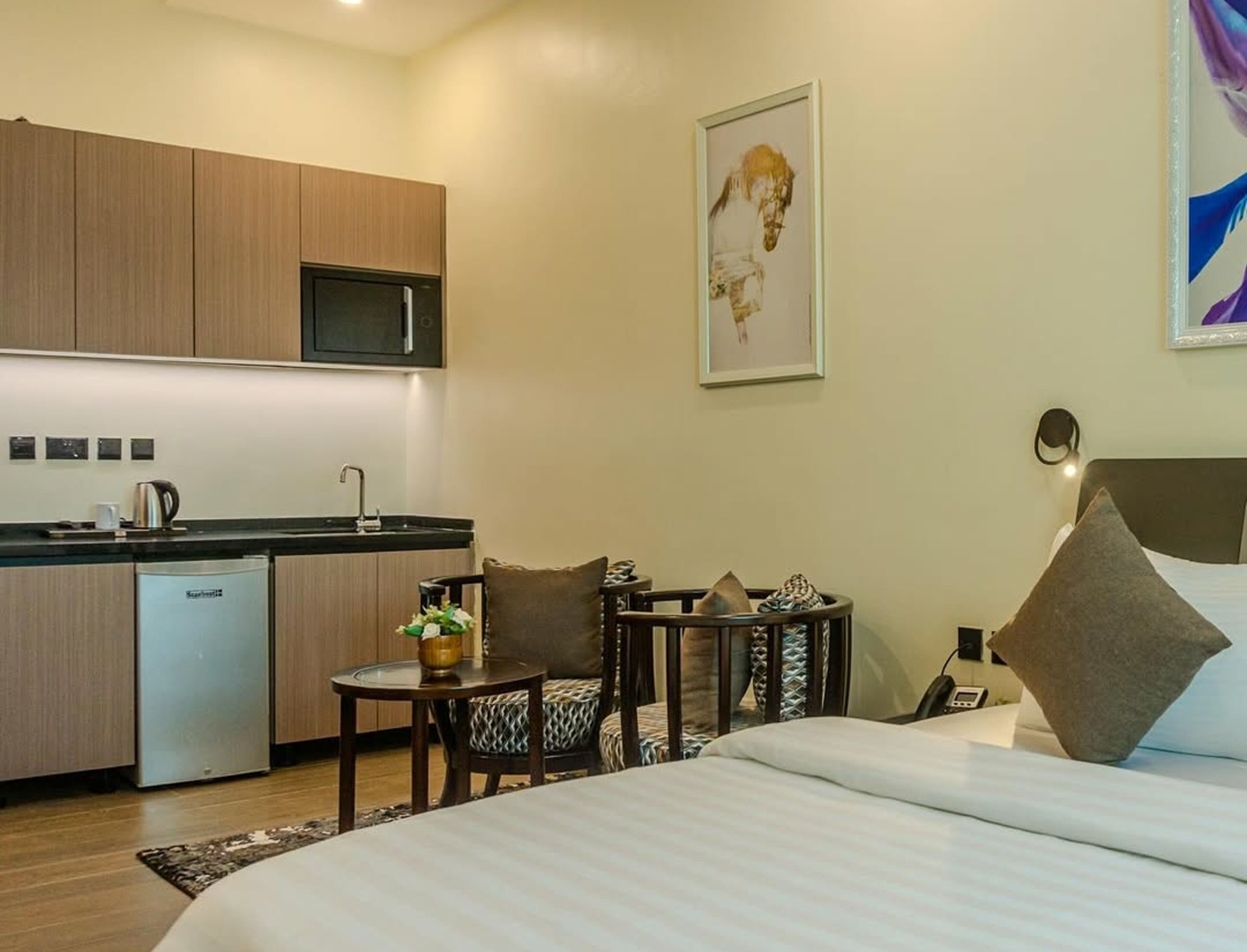 A modern hotel room with a bed, a small table with chairs, a kitchen area with a microwave, and a painting on the wall.