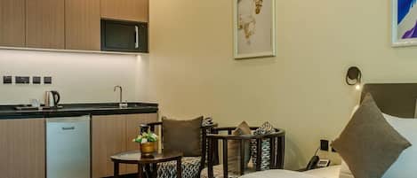 A modern hotel room with a bed, a small table with chairs, a kitchen area with a microwave, and a painting on the wall.