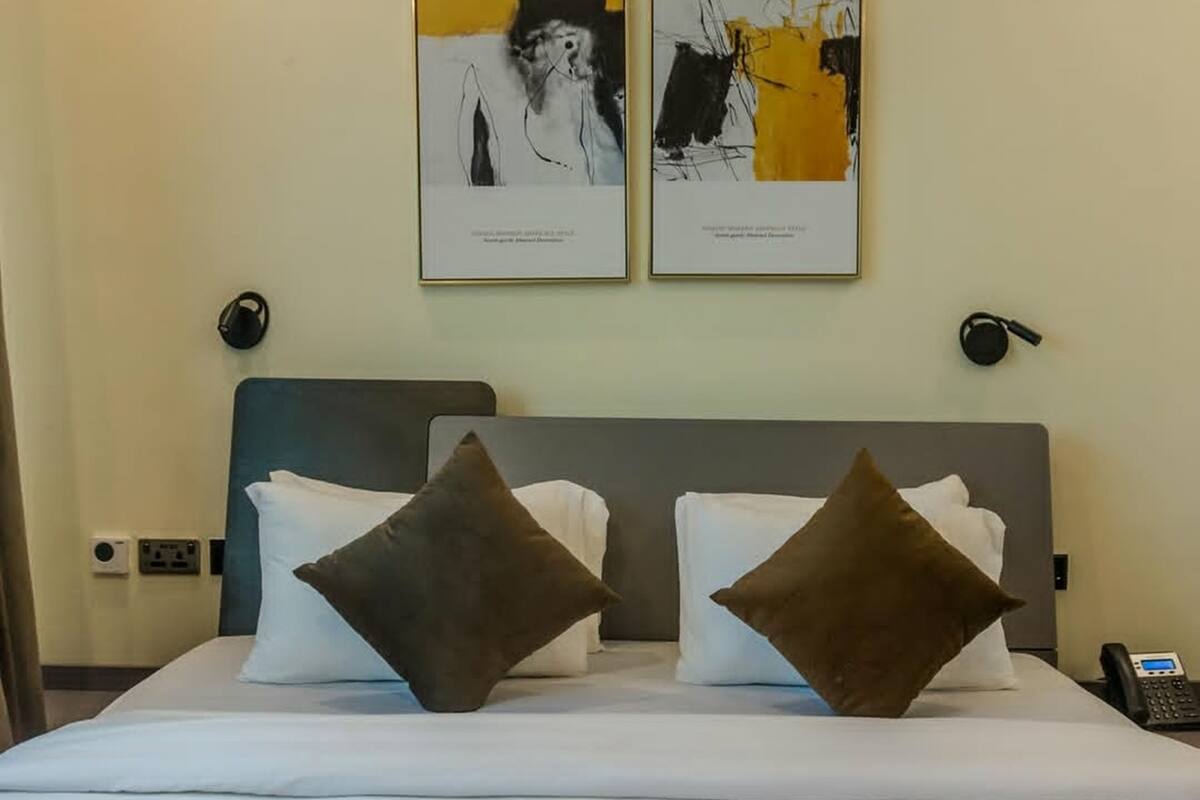 A hotel room with a bed, two pillows, a headboard, a wall-mounted phone, and two framed artworks above the bed.