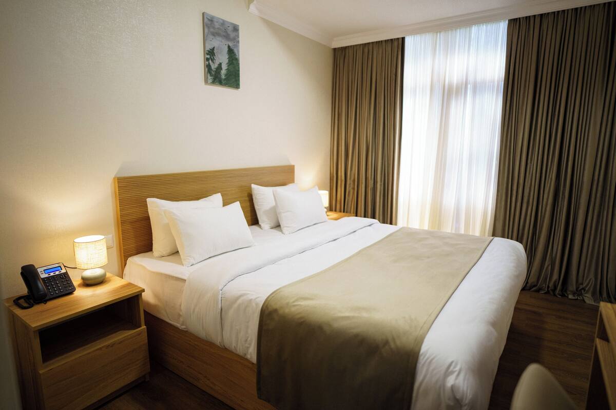 Een hotelkamer met een houten bed, witte beddengoed, een bedlampje, een telefoon op het nachtkastje, en een raam met gordijnen.