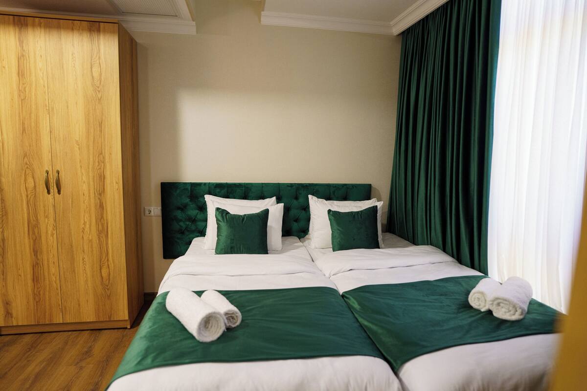 Een hotelkamer met een tweepersoonsbed, groen beddengoed, een houten garderobe en een groot raam met gordijnen.