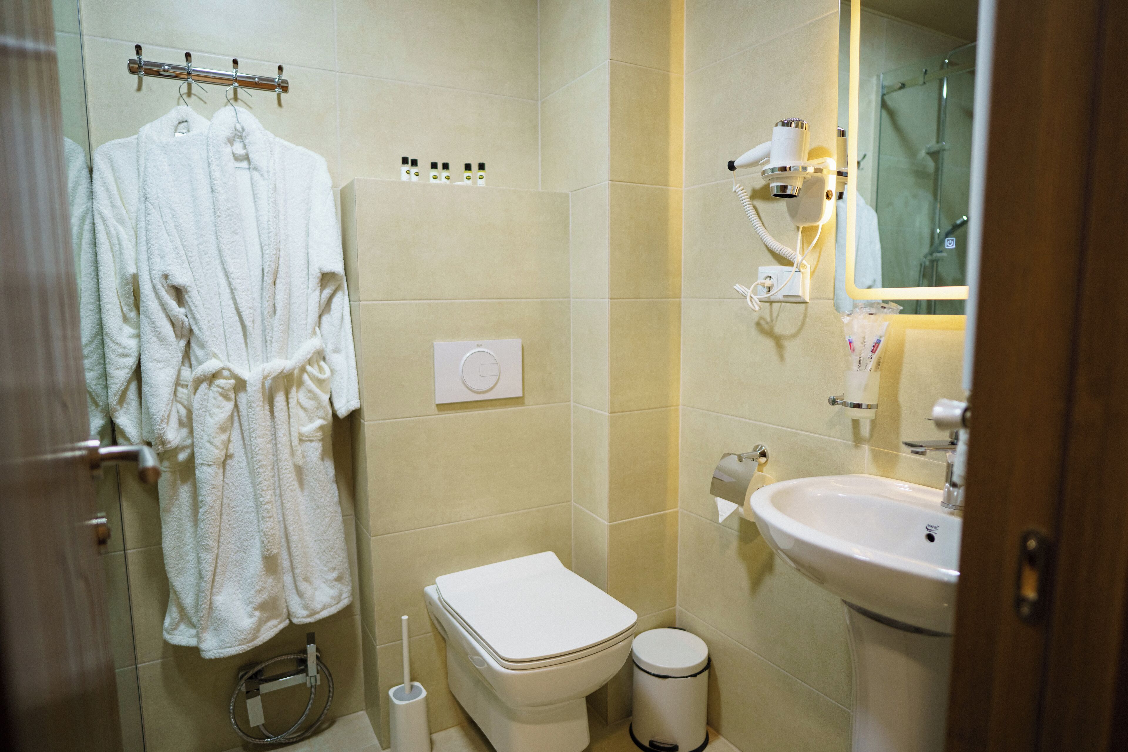 Un baño con inodoro, lavamanos y espejo. Hay toallas colgadas en un perchero y un cubo de basura.