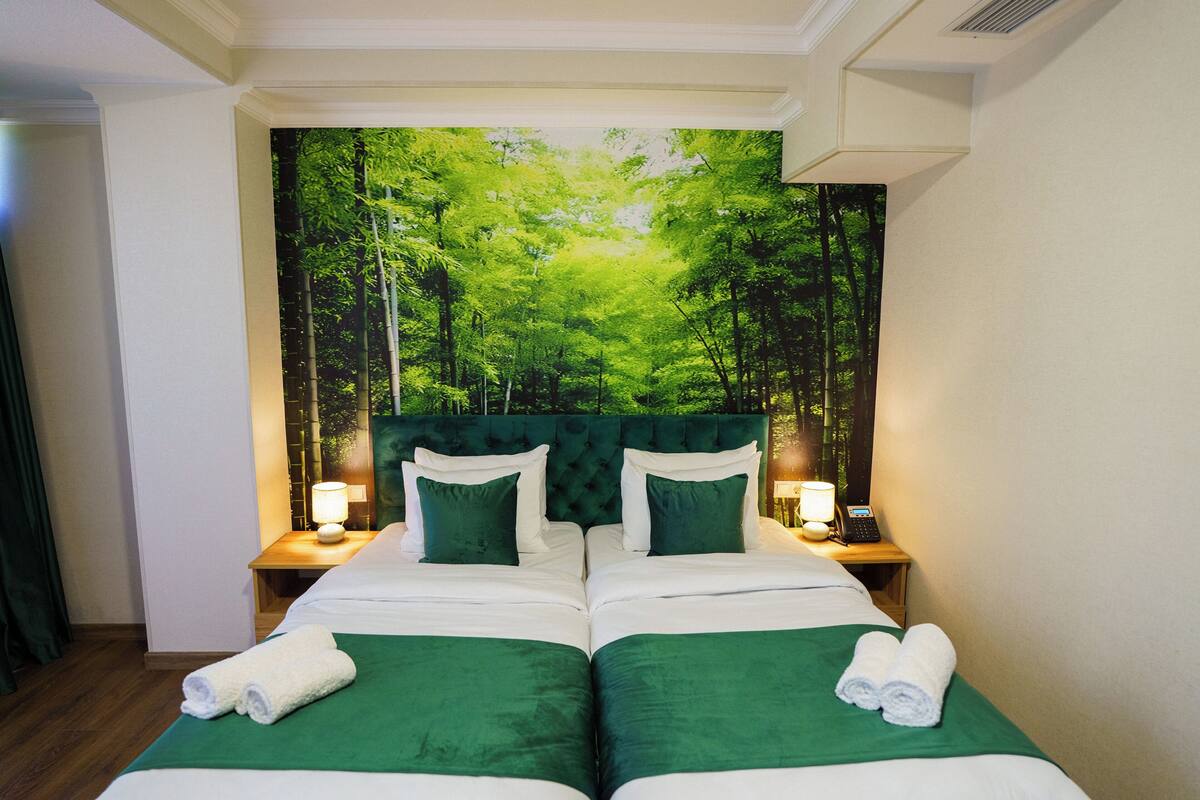 Een tweepersoonsbed met groene beddengoed en twee kussens, met aan beide zijden nachtkastjes met lampen, in een kamer met een bosdecoratie op de muur.