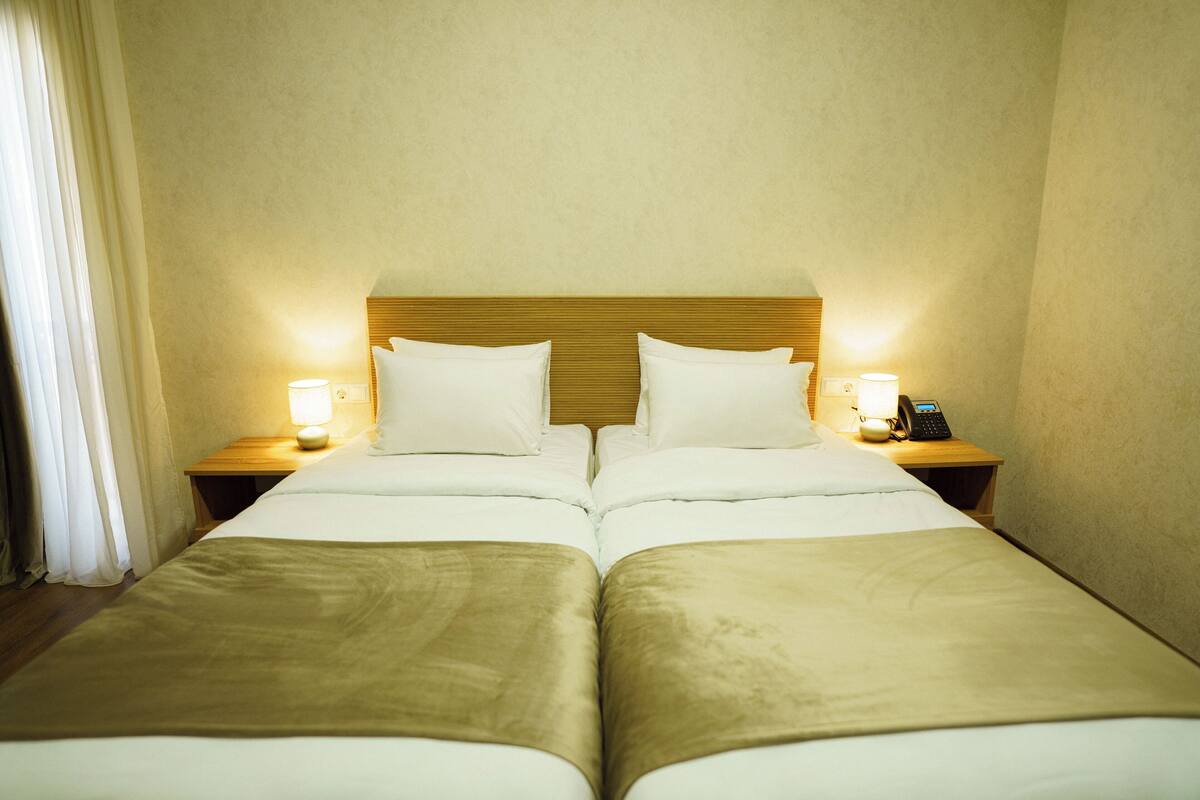 Een tweepersoonsbed met een houten hoofdeinde, twee nachtkastjes met lampen, en een gordijn aan de linkerkant.