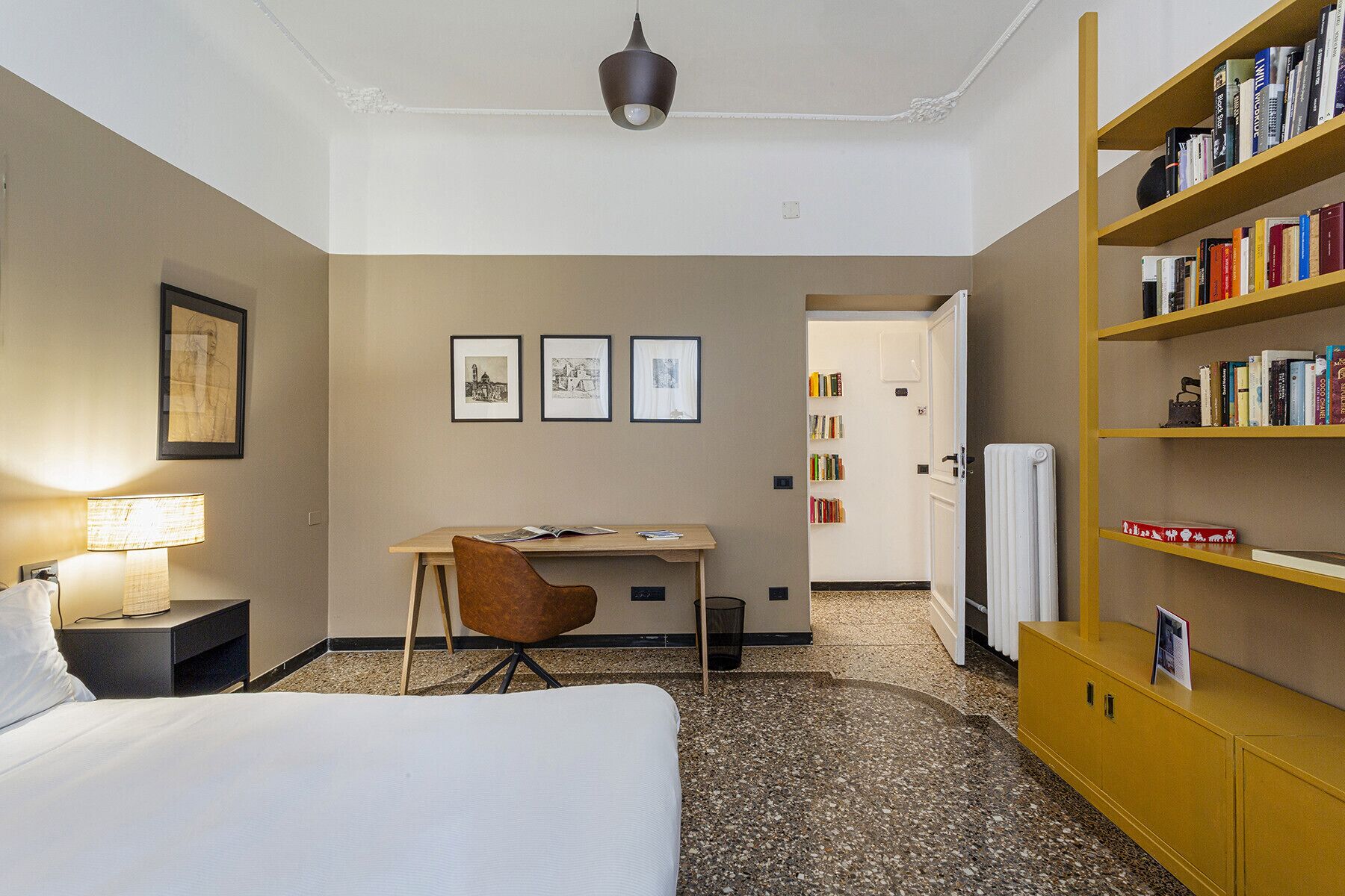 Quarto de hotel com cama, escrivaninha, cadeira, estante e quadros na parede.