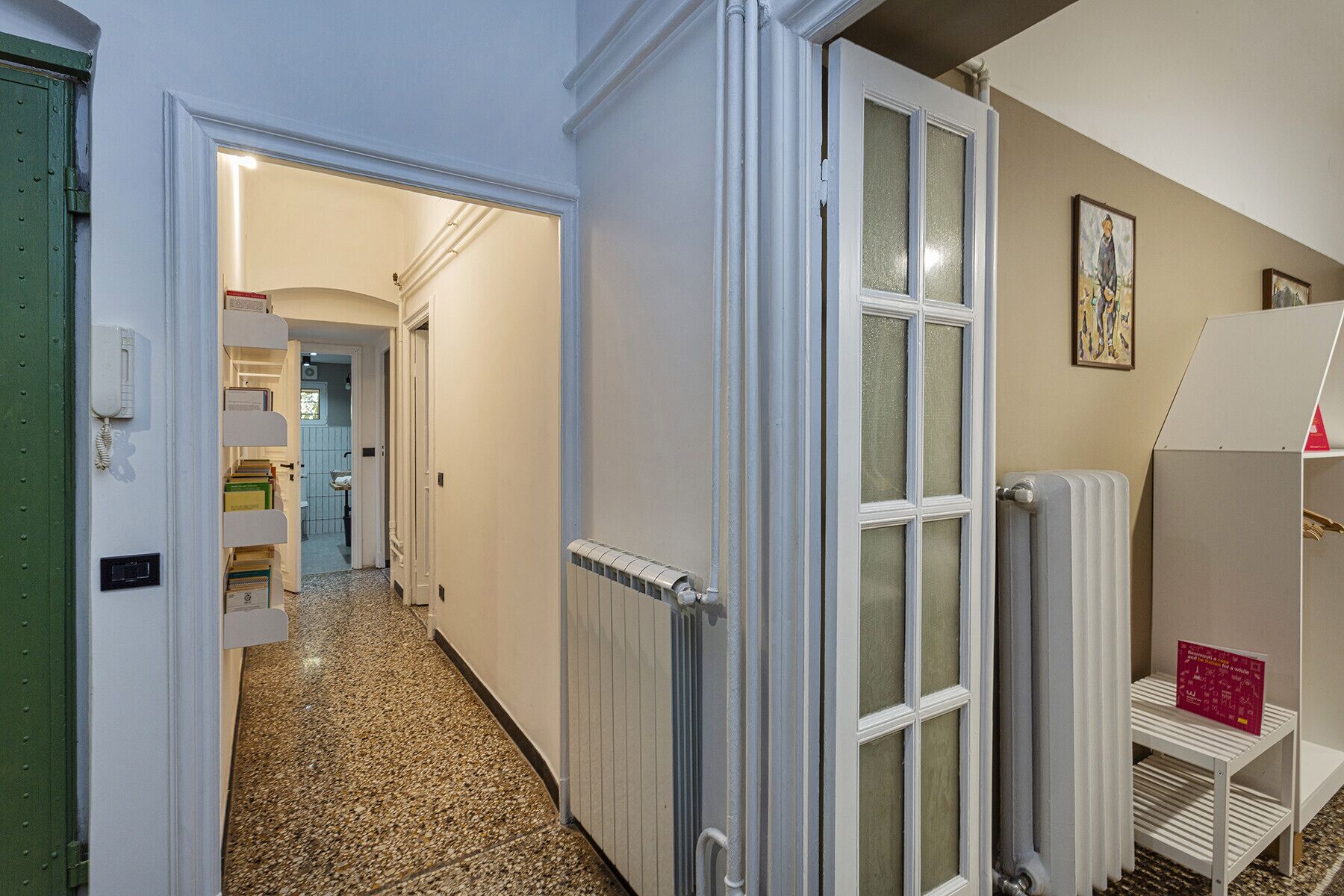 Un pasillo con un radiador, una puerta que da al baño y una estantería con libros.