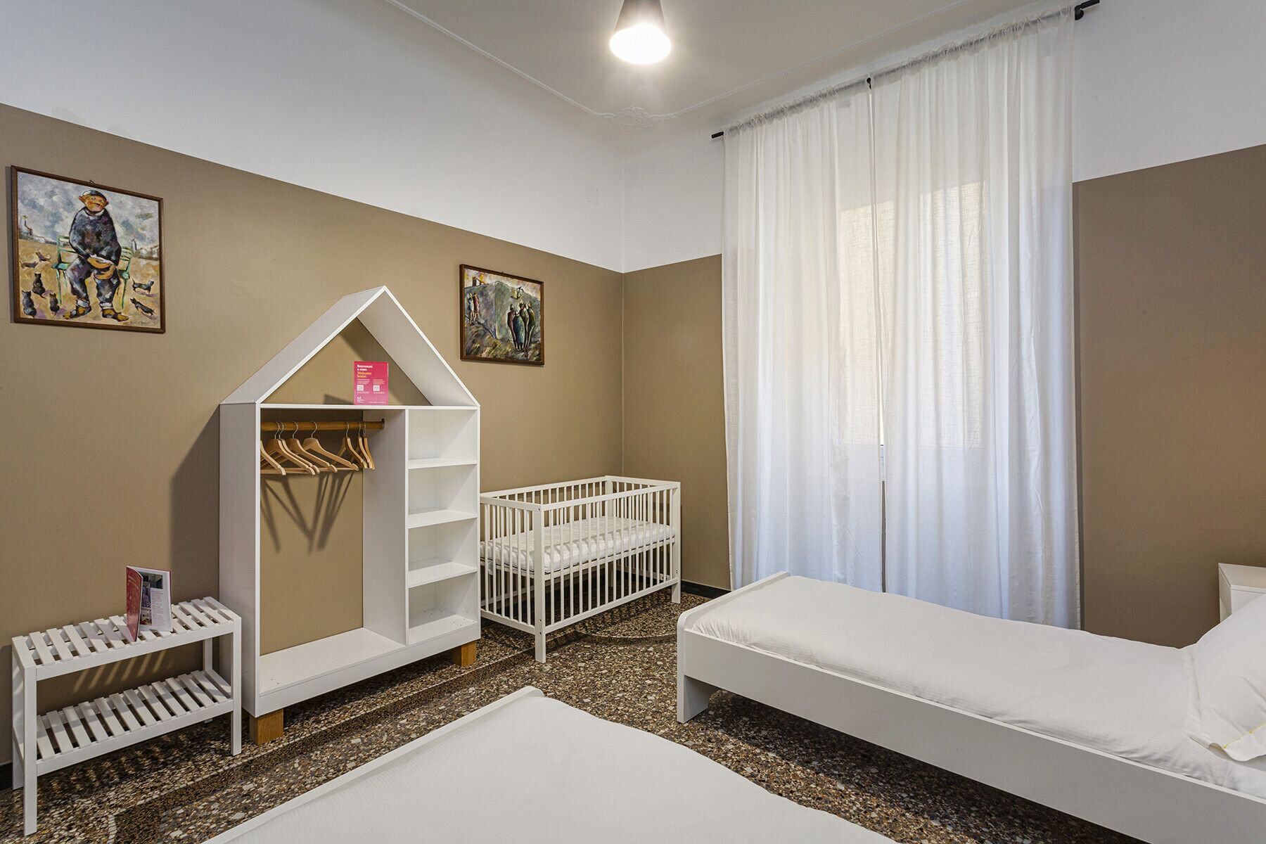 A room with a crib, a small bed, a white playhouse, and two paintings on the wall.